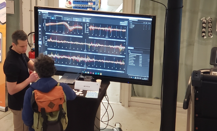 The Moveck stand at the SOFAMEA 2024 congress in Nantes: multi-trial kinematic curves on a large display while a team member talks a visitor through the analysis